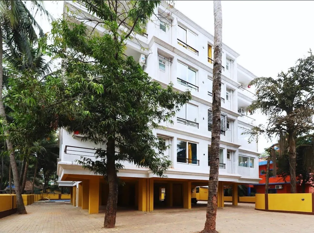 White four-storey apartment building framed by palm and mango trees in Goa